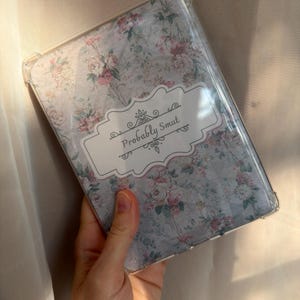 Könnte beinhalten: Eine transparente Schutzhülle für E-Reader mit hellblauem Hintergrund und einem Blumenmuster aus rosa und weißen Rosen. In der Mitte befindet sich ein weißes rechteckiges Etikett mit dekorativen Schnörkeln und dem Text Probably Smut.
