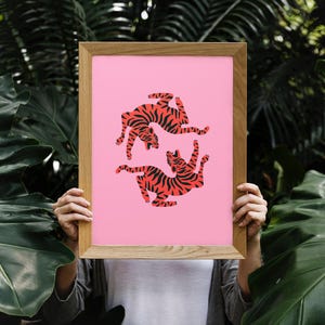 K&ouml;nnte beinhalten: Ein gerahmter Kunstdruck mit zwei stilisierten Tigern in rot-schwarzen Streifen auf rosa Hintergrund. Das Kunstwerk befindet sich in einem Holzrahmen, vor einem Hintergrund aus &uuml;ppigem gr&uuml;nem Laub.