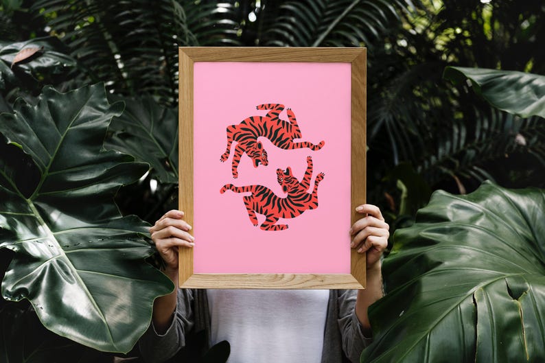 K&ouml;nnte beinhalten: Ein gerahmter Kunstdruck mit zwei stilisierten Tigern in rot-schwarzen Streifen auf rosa Hintergrund. Das Kunstwerk befindet sich in einem Holzrahmen, vor einem Hintergrund aus &uuml;ppigem gr&uuml;nem Laub.