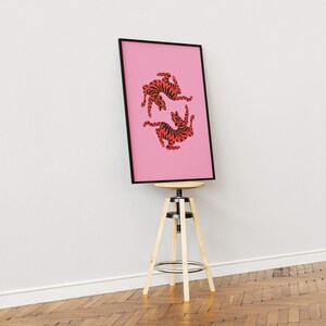 K&ouml;nnte beinhalten: Ein gerahmter rosa Kunstdruck mit zwei stilisierten orange-schwarzen Tigerillustrationen. Das Kunstwerk wird auf einem Holzschemel mit schwarzem Rahmen vor einer wei&szlig;en Wand und einem Holzboden pr&auml;sentiert.