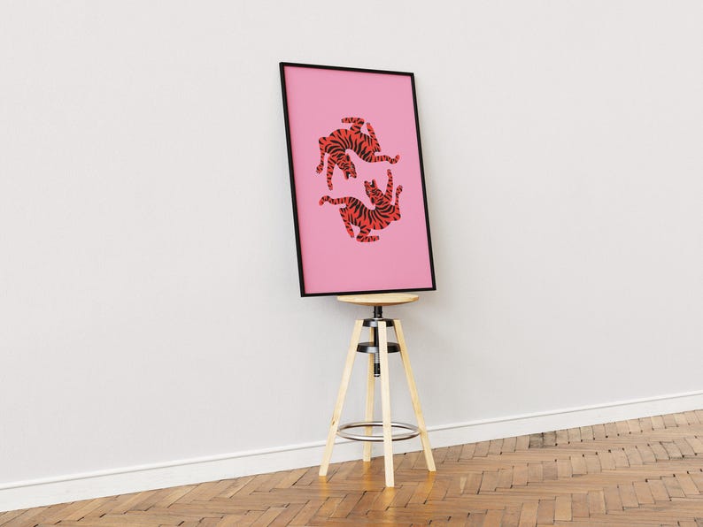 K&ouml;nnte beinhalten: Ein gerahmter rosa Kunstdruck mit zwei stilisierten orange-schwarzen Tigerillustrationen. Das Kunstwerk wird auf einem Holzschemel mit schwarzem Rahmen vor einer wei&szlig;en Wand und einem Holzboden pr&auml;sentiert.