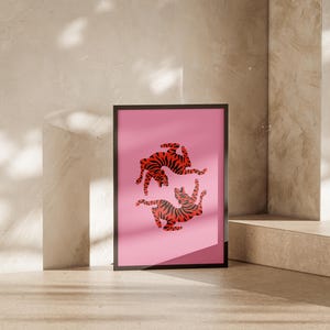 K&ouml;nnte beinhalten: Ein gerahmter Kunstdruck mit einem rosa Hintergrund, der zwei stilisierte Tiger mit rot-schwarzen Streifen zeigt. Das Kunstwerk wird in einem schwarzen Rahmen pr&auml;sentiert, der an einer neutral get&ouml;nten Wand lehnt.