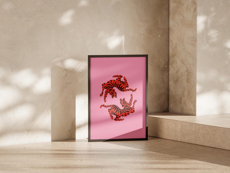 K&ouml;nnte beinhalten: Ein gerahmter Kunstdruck mit einem rosa Hintergrund, der zwei stilisierte Tiger mit rot-schwarzen Streifen zeigt. Das Kunstwerk wird in einem schwarzen Rahmen pr&auml;sentiert, der an einer neutral get&ouml;nten Wand lehnt.