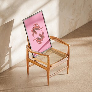 K&ouml;nnte beinhalten: Ein gerahmter rosa Kunstdruck mit zwei orangefarbenen und schwarzen Tigerillustrationen. Das Kunstwerk lehnt an einem Holzstuhl mit einem gewebten Sitz. Der Stuhl steht auf einer neutralen Oberfl&auml;che, mit Schatten. Der Stuhl ist aus Holz.