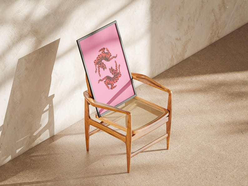 K&ouml;nnte beinhalten: Ein gerahmter rosa Kunstdruck mit zwei orangefarbenen und schwarzen Tigerillustrationen. Das Kunstwerk lehnt an einem Holzstuhl mit einem gewebten Sitz. Der Stuhl steht auf einer neutralen Oberfl&auml;che, mit Schatten. Der Stuhl ist aus Holz.