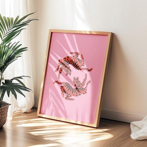 K&ouml;nnte beinhalten: Ein gerahmter rosa Kunstdruck mit zwei stilisierten Tigern in orangefarbenen und grauen Streifen. Das Kunstwerk wird in einem hellen Holzrahmen pr&auml;sentiert, der an einer Wand lehnt. Ein Topfpflanze und Vorh&auml;nge befinden sich im Hintergrund.