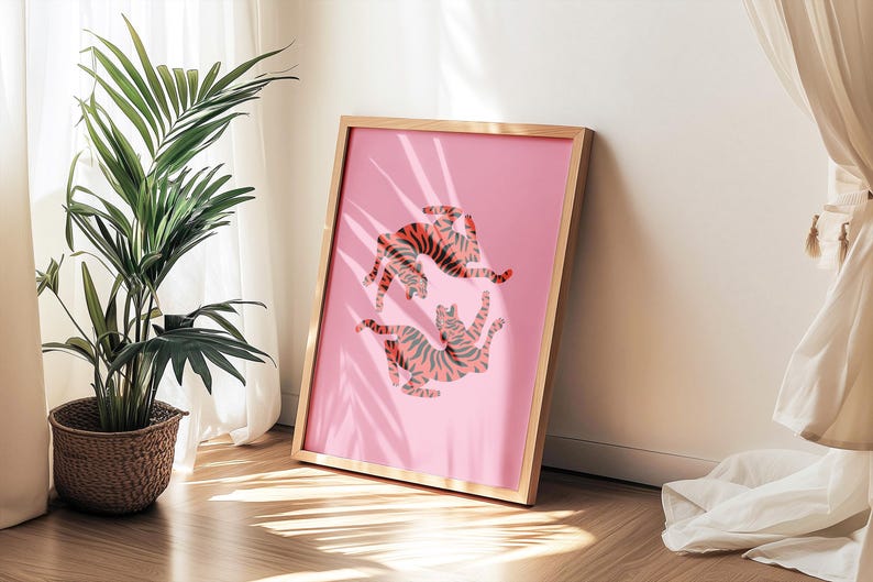 K&ouml;nnte beinhalten: Ein gerahmter rosa Kunstdruck mit zwei stilisierten Tigern in orangefarbenen und grauen Streifen. Das Kunstwerk wird in einem hellen Holzrahmen pr&auml;sentiert, der an einer Wand lehnt. Ein Topfpflanze und Vorh&auml;nge befinden sich im Hintergrund.