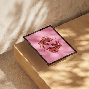 K&ouml;nnte beinhalten: Ein gerahmter Kunstdruck mit zwei stilisierten Tigern in gespiegelter Pose auf rosa Hintergrund. Die Tiger sind orange mit schwarzen Streifen. Der Rahmen ist schwarz, und der Druck wird auf einer Holzoberfl&auml;che pr&auml;sentiert.