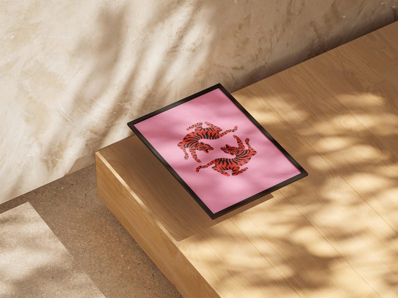 K&ouml;nnte beinhalten: Ein gerahmter Kunstdruck mit zwei stilisierten Tigern in gespiegelter Pose auf rosa Hintergrund. Die Tiger sind orange mit schwarzen Streifen. Der Rahmen ist schwarz, und der Druck wird auf einer Holzoberfl&auml;che pr&auml;sentiert.