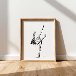 Puede incluir: Dibujo lineal en blanco y negro enmarcado de una bailarina en una pose elegante. La obra de arte está en un marco de madera clara y se apoya contra una pared blanca sobre un suelo de madera. La bailarina está representada a medio salto, con una pierna extendida.