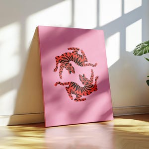 K&ouml;nnte beinhalten: Ein pinkfarbener Leinwanddruck mit zwei roten Tigern mit schwarzen Streifen in einer dynamischen Pose. Das Kunstwerk steht vor einer Wand in einem gut beleuchteten Raum. Der Hintergrund ist einfarbig pink.