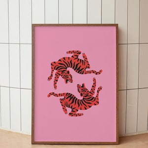 K&ouml;nnte beinhalten: Ein gerahmter Kunstdruck mit einem rosa Hintergrund, der drei stilisierte Tiger in Rot und Schwarz zeigt. Die Tiger sind in dynamischen Posen dargestellt. Der Rahmen hat eine warme braune Farbe und tr&auml;gt zur Gesamt&auml;sthetik des Kunstwerks bei.