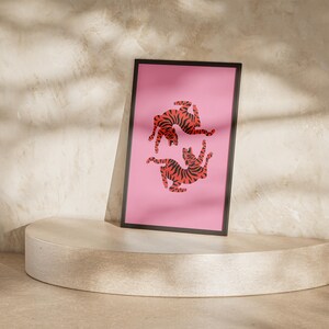 K&ouml;nnte beinhalten: Ein gerahmter Kunstdruck mit einem rosa Hintergrund, der zwei stilisierte Tiger mit orangefarbenen und schwarzen Streifen zeigt. Das Kunstwerk wird auf einem runden, hellen Sockel pr&auml;sentiert. Der Hintergrund ist eine strukturierte Wand.
