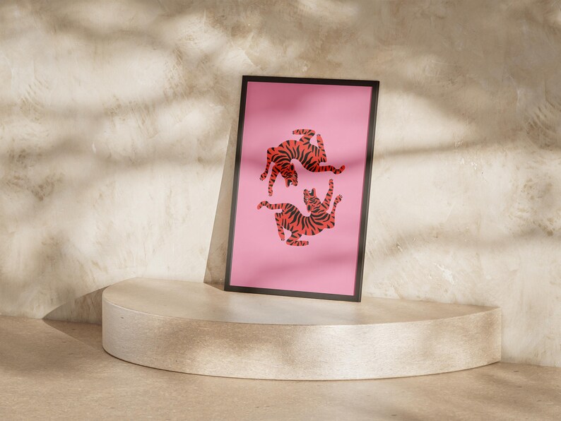 K&ouml;nnte beinhalten: Ein gerahmter Kunstdruck mit einem rosa Hintergrund, der zwei stilisierte Tiger mit orangefarbenen und schwarzen Streifen zeigt. Das Kunstwerk wird auf einem runden, hellen Sockel pr&auml;sentiert. Der Hintergrund ist eine strukturierte Wand.