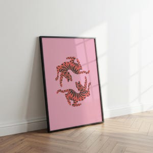 K&ouml;nnte beinhalten: Ein gerahmter Kunstdruck mit einem rosa Hintergrund, der zwei Tiger mit orangefarbenen und schwarzen Streifen zeigt. Die Tiger sind in dynamischen Posen dargestellt. Der Druck ist in einem schwarzen Rahmen gefasst. Das Kunstwerk lehnt an einer wei&szlig;en Wand auf einem Holzboden.
