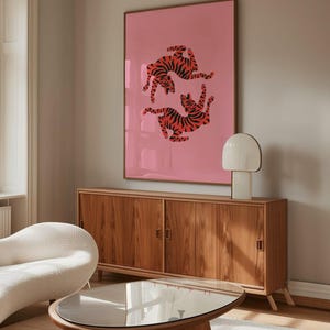 K&ouml;nnte beinhalten: Ein gerahmter rosa Kunstdruck mit zwei stilisierten Tigern mit orangefarbenen und schwarzen Streifen. Das Kunstwerk h&auml;ngt &uuml;ber einem Holzschrank mit zwei T&uuml;ren und einem Couchtisch mit Glasplatte. Eine wei&szlig;e Lampe und ein geschwungenes wei&szlig;es Sofa sind ebenfalls zu sehen.