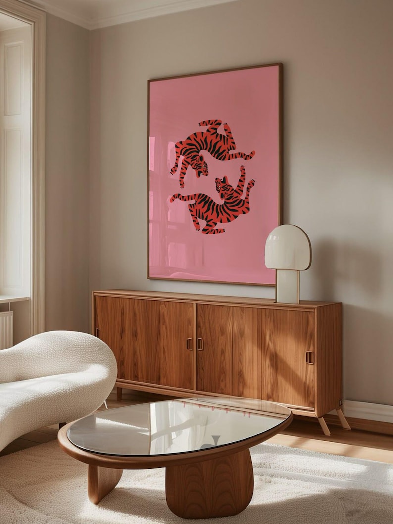 K&ouml;nnte beinhalten: Ein gerahmter rosa Kunstdruck mit zwei stilisierten Tigern mit orangefarbenen und schwarzen Streifen. Das Kunstwerk h&auml;ngt &uuml;ber einem Holzschrank mit zwei T&uuml;ren und einem Couchtisch mit Glasplatte. Eine wei&szlig;e Lampe und ein geschwungenes wei&szlig;es Sofa sind ebenfalls zu sehen.