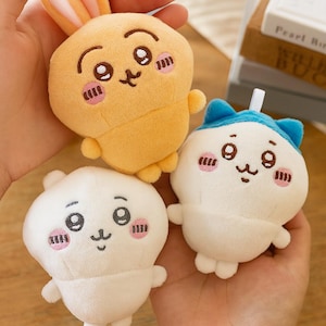 May include: Three plush toys with cartoon faces are held in a hand. The toys include a yellow bunny, a white bear, and a white cat with a blue hat. Each toy has a smiling face with pink cheeks and black details.