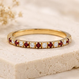 May include: A gold band ring with alternating red and white gemstones. The ring is set on a light-colored stone surface, with dried flowers in the background. The gemstones are evenly spaced around the band.