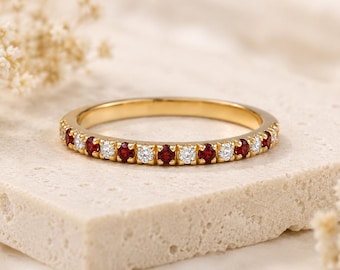 Può includere: Un anello a fascia dorato con pietre preziose rosse e bianche alternate. L'anello è posto su una superficie di pietra chiara, con fiori secchi sullo sfondo. Le pietre preziose sono distanziate uniformemente.