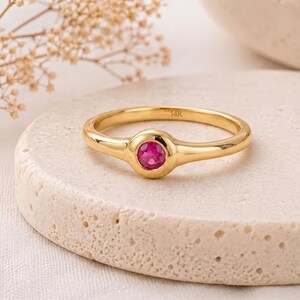 May include: A gold ring with a round, bright pink gemstone set in the center. The ring is on a textured, off-white surface. The ring band is thin and smooth, with a slight curve on either side of the gemstone.