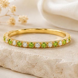 May include: A gold band ring with alternating green and white gemstones. The ring is set on a light-colored stone surface, with a white cloth and dried flowers in the background. The ring is a delicate piece of jewelry.