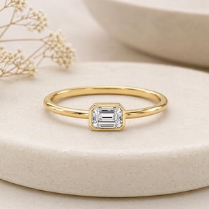 May include: A gold ring with a rectangular, emerald-cut diamond center stone. The ring has a thin, polished band and a bezel setting. The ring is displayed on a light-colored, textured surface, with a blurred background.