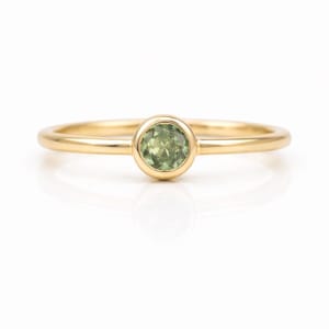 May include: A gold ring featuring a round, light green gemstone. The ring has a thin, polished band and a bezel setting that holds the gemstone securely. The ring is displayed against a white background.