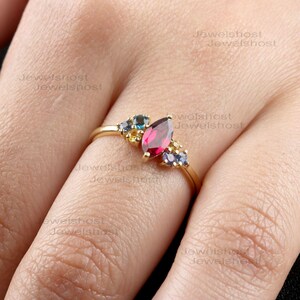 May include: A gold ring featuring a marquise-cut, deep red gemstone as the focal point, flanked by clusters of smaller blue, yellow, and purple gemstones. The ring's band is a thin, polished gold.