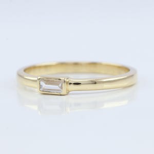 以下が含まれることがあります： 中央に長方形の透明な宝石をあしらったゴールドリング。バンドは滑らかで磨かれており、光を反射します。宝石は水平にセットされており、シンプルなデザインにエレガントさを加えています。