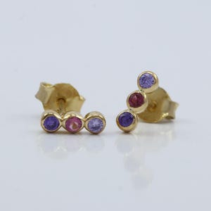 May include: A pair of gold stud earrings, each featuring three colorful gemstones. One earring has purple, pink, and purple stones, while the other has purple, red, and purple stones. The earrings are set against a white background.