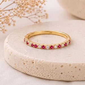 Peut inclure: Une bague en or avec des pierres précieuses rouges et claires alternées. La bague est posée sur une surface claire et texturée. La bague est marquée "14K".