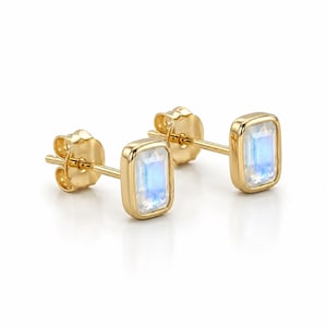 May include: A pair of gold stud earrings featuring rectangular, faceted moonstones set in smooth gold bezels. Each earring includes a gold post and a matching gold butterfly clutch backing, showcasing the iridescent blue glow of the stones.