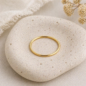 May include: A thin, gold-colored ring rests on a light-colored, textured stone. The ring is circular and appears to be made of metal. The stone is oval-shaped and has a natural, organic appearance. Dried flowers and a linen cloth are in the background.