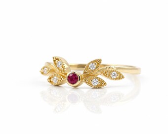 Könnte beinhalten: Ein Goldring mit einem zentralen, runden, roten Rubin in einer Zargenfassung, flankiert von blattförmigen Akzenten. Jedes Blatt ist mit kleinen, klaren, rund geschliffenen Diamanten in Gold mit Milgrain-Rand verziert, was ein symmetrisches Design ergibt.