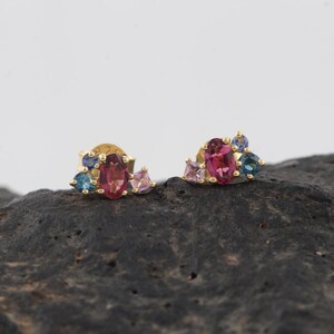 May include: A pair of gold stud earrings, each featuring a central oval-cut pink gemstone surrounded by smaller round and square-cut blue and light purple stones in a cluster arrangement, set in a yellow gold metal base.