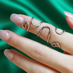 Op de afbeelding: Een zilverkleurige ring voor meerdere vingers met een uniek, vloeiend ontwerp. De ring wikkelt zich om de vinger met gebogen en geluste elementen, waardoor een moderne en artistieke uitstraling ontstaat. De sieraden zijn geplaatst tegen een groene achtergrond.