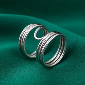 May include: A silver-colored ring with a unique design. The ring features multiple bands of metal that curve and loop, creating an open, eye-catching shape. The ring is set against a green background.