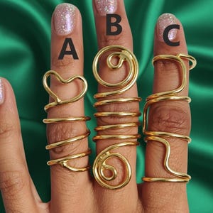 May include: Gold-colored rings in various designs, including a heart, a spiral, and a geometric shape. The rings are worn on fingers, showcasing their intricate wire-like structures. The background is a solid green color.