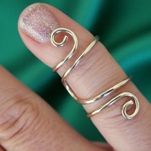 Può includere: Un anello da dito color oro con un design unico a spirale. L'anello si avvolge attorno al dito con due sezioni curve e un vortice decorativo a ciascuna estremità. L'anello è indossato su un dito con smalto glitterato.