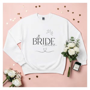 May include: White sweatshirt with the text "BRIDE babe" in black script. The design includes a ring, doves, and a heart. The sweatshirt is displayed on a pink surface with gold confetti and a bouquet of white roses.