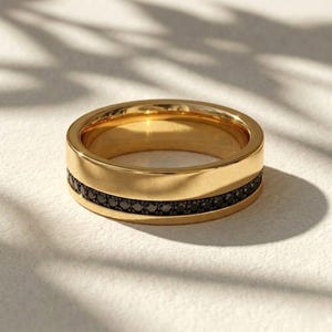 May include: A gold ring featuring a row of small, black gemstones. The ring has a flat, wide design with a polished finish. The gemstones are set in a band around the centre of the ring, creating a contrasting detail.