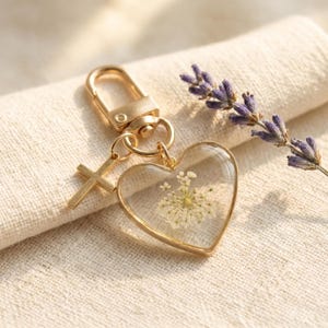 May include: A gold-coloured keyring featuring a heart-shaped pendant with dried white flowers. The pendant is framed in gold and hangs with a small gold cross charm. A gold clasp and a sprig of lavender are also visible.