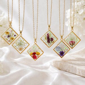 Puede incluir: Seis collares dorados con colgantes en forma de diamante. Cada colgante contiene resina transparente que encierra varias flores secas prensadas, incluyendo flores amarillas, rojas, moradas y blancas. Cuelgan de cadenas de metal fino dorado.