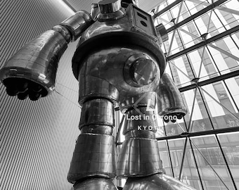 Könnte beinhalten: Eine große Metallroboter-Skulptur mit genieteten Paneelen und zylindrischen Gliedmaßen. Der Roboter steht in einem modernen Gebäude mit Glasfenstern und dem Text "Lost in Orono Kyoto". Die Skulptur ist in Schwarzweiß gehalten.