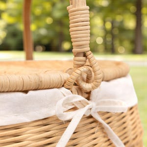 May include: Close-up of a wicker picnic basket with a woven handle and a white fabric lining. The handle is made of tightly wound, light-colored material. A white ribbon is tied in a bow on the front of the basket.