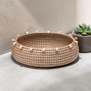 May include: A round, woven basket with a natural beige color and a series of small, light-colored wooden beads around the rim. The basket is sitting on a gray surface, with a succulent plant in a gray pot in the background.