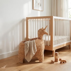 May include: A woven wicker unicorn-shaped storage basket with a tan blanket inside. A wooden crib with white slats is in the background. Wooden blocks and a fox figurine are on the floor. The room has a neutral color palette.