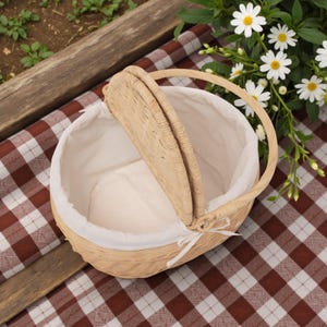 May include: A light brown wicker picnic basket with a white fabric lining. The basket has a handle and a hinged lid. It sits on a red and white checkered picnic blanket, with flowers in the background.