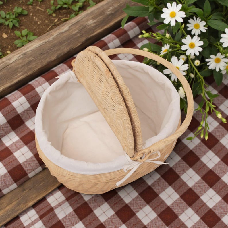 May include: A light brown wicker picnic basket with a white fabric lining. The basket has a handle and a hinged lid. It sits on a red and white checkered picnic blanket, with flowers in the background.